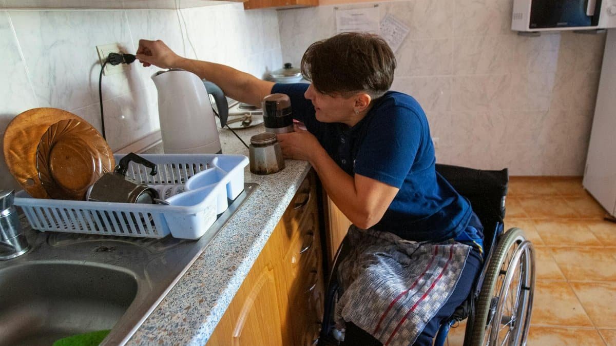 A woman in a wheelchair prepares coffee in a cozy kitchen setting.
