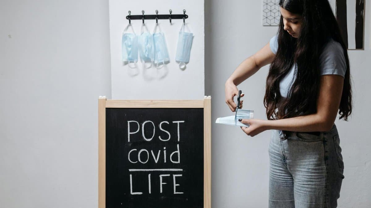 A young woman cutting a face mask next to a chalkboard reading 'POST covid LIFE' indoors.