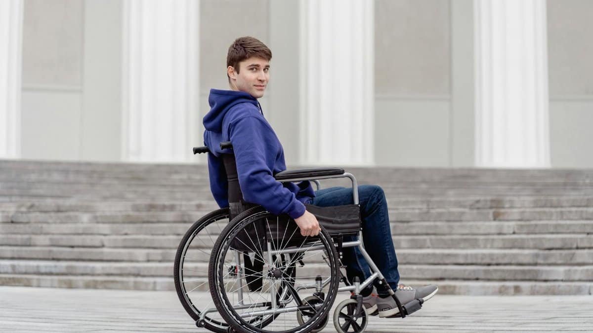 Young adult man in a wheelchair wearing a hoodie, seated outdoors by steps, looking over his shoulder.