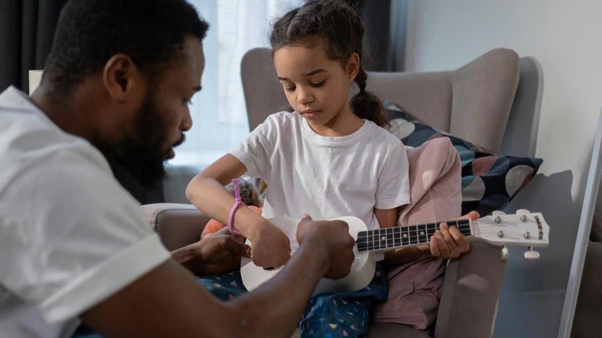 A father and daughter bonding indoors while learning to play the ukulele, creating a warm family moment.