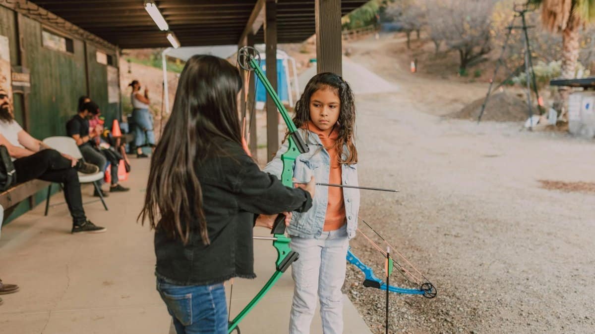 Two young girls practice archery at an outdoor range, learning to use a bow and arrow.