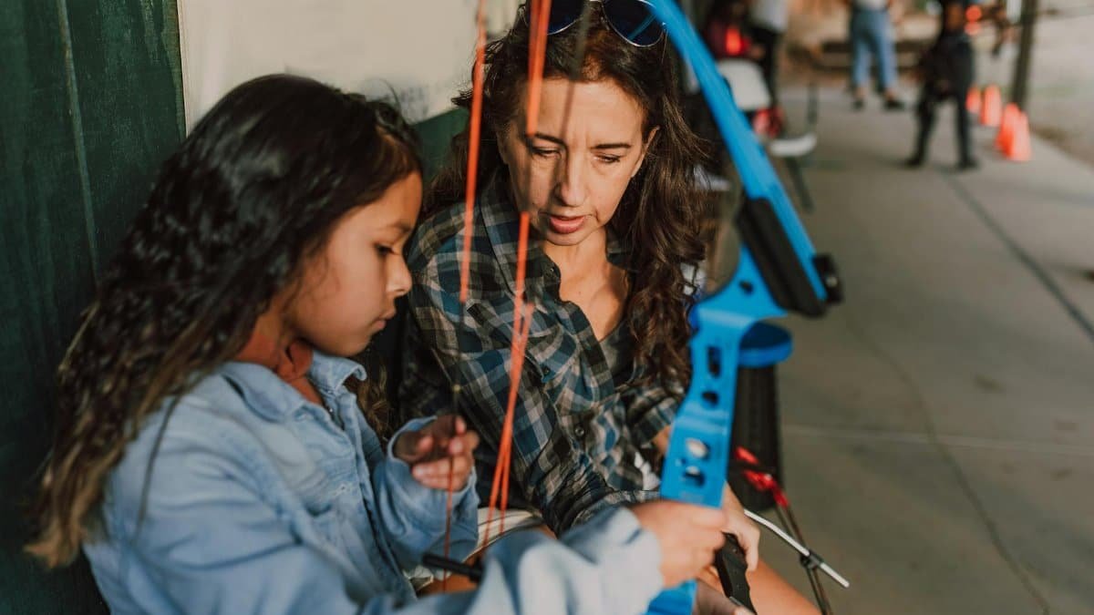 A woman instructs a young girl on archery techniques, holding a blue bow outdoors.