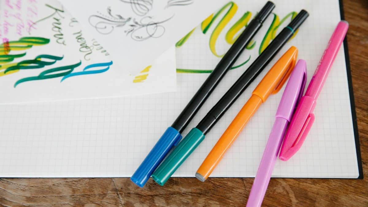 Colorful pens and calligraphy sheets on a wooden table, perfect for art and crafts enthusiasts.