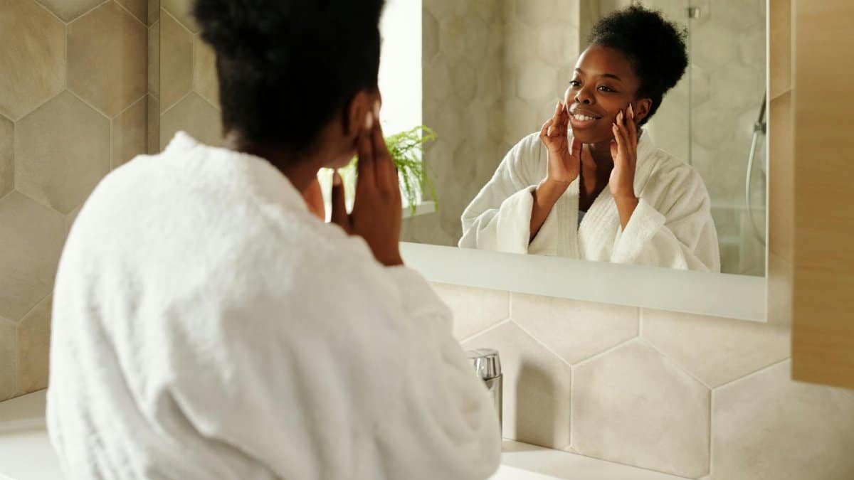 A woman in a bathrobe smiles at her reflection in a modern bathroom mirror.