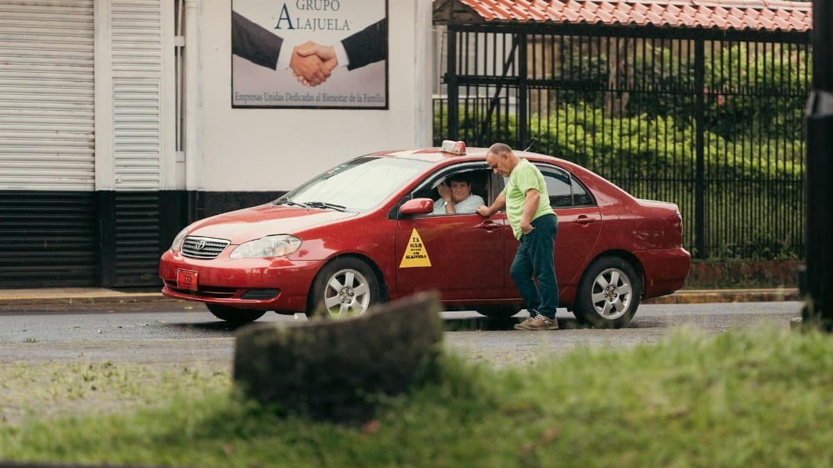 Two people interacting outside a red taxi on a rainy street in Alajuela, Costa Rica.