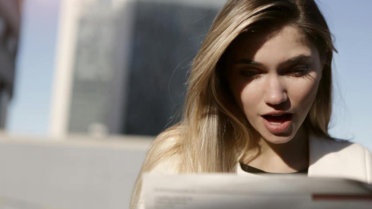 A young woman with blonde hair expresses surprise while reading a newspaper outdoors in daylight.