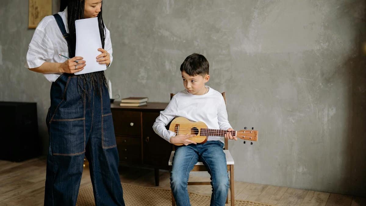 Boy with ukulele receiving instructions from teacher in a cozy classroom setting.