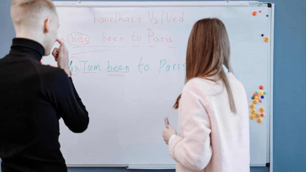 Two students studying English grammar at a whiteboard in a classroom setting.
