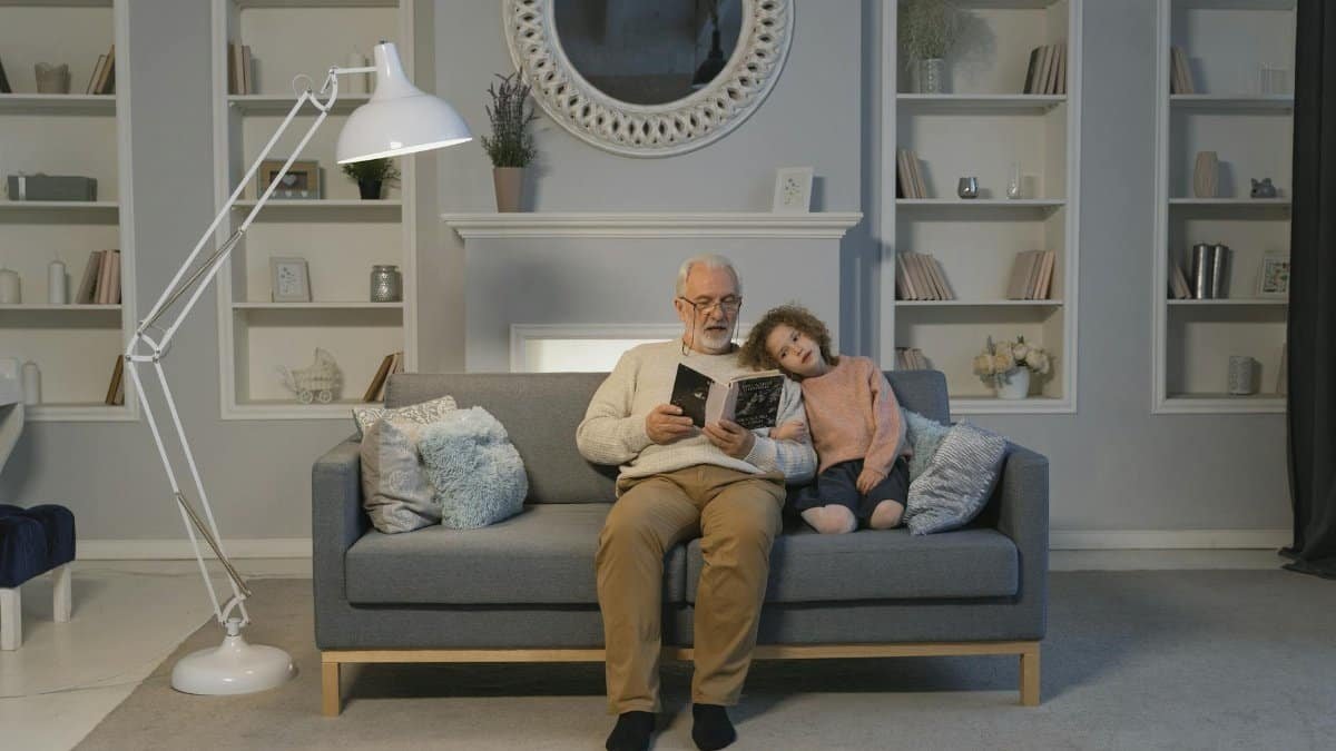 A cozy scene with a grandfather reading to his granddaughter at home on the sofa.