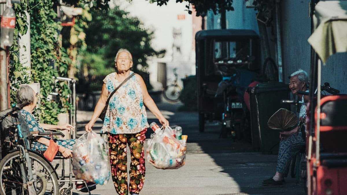 An elderly East Asian woman walks through a Beijing street carrying recyclables, showcasing urban life.