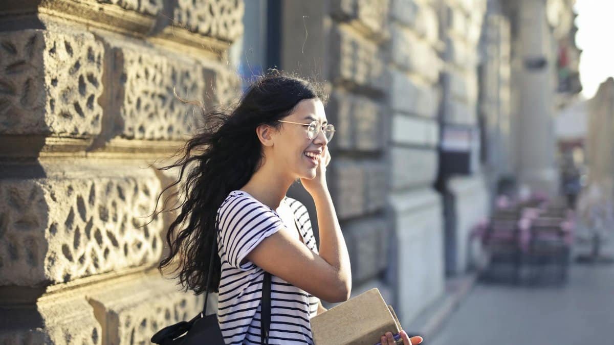 A smiling woman with flowing hair enjoys a windy day, holding a book against an urban backdrop.
