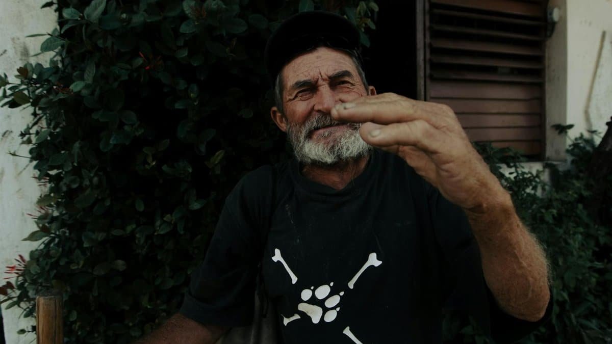 An elderly bearded man stands outdoors, gesturing with a relaxed expression in a casual setting.