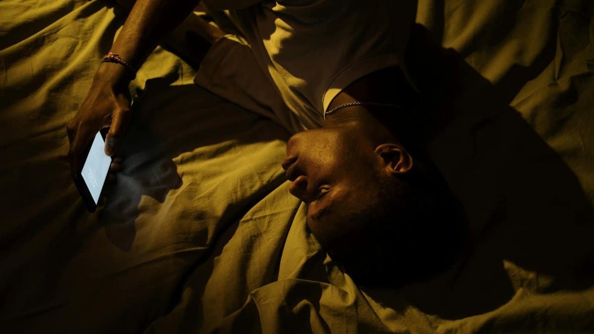 A man lying in bed at night, illuminated by a smartphone, suggesting insomnia or sleeplessness.