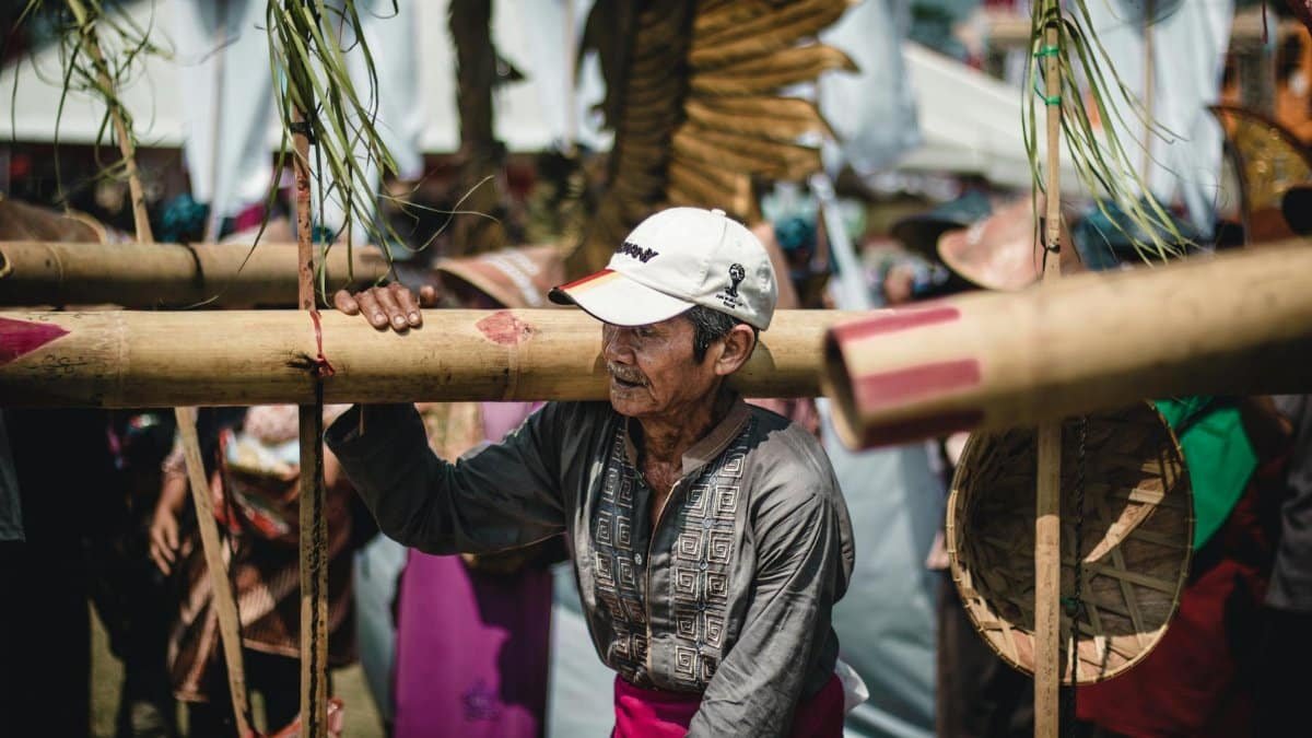Serious mature Asian male in casual clothes walking with long bamboo stem on shoulder in local village