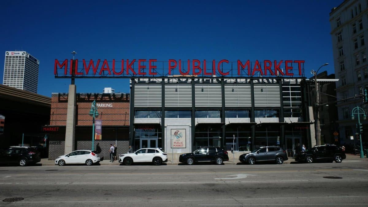A vibrant street view of Milwaukee Public Market with cars and pedestrians under a clear blue sky.