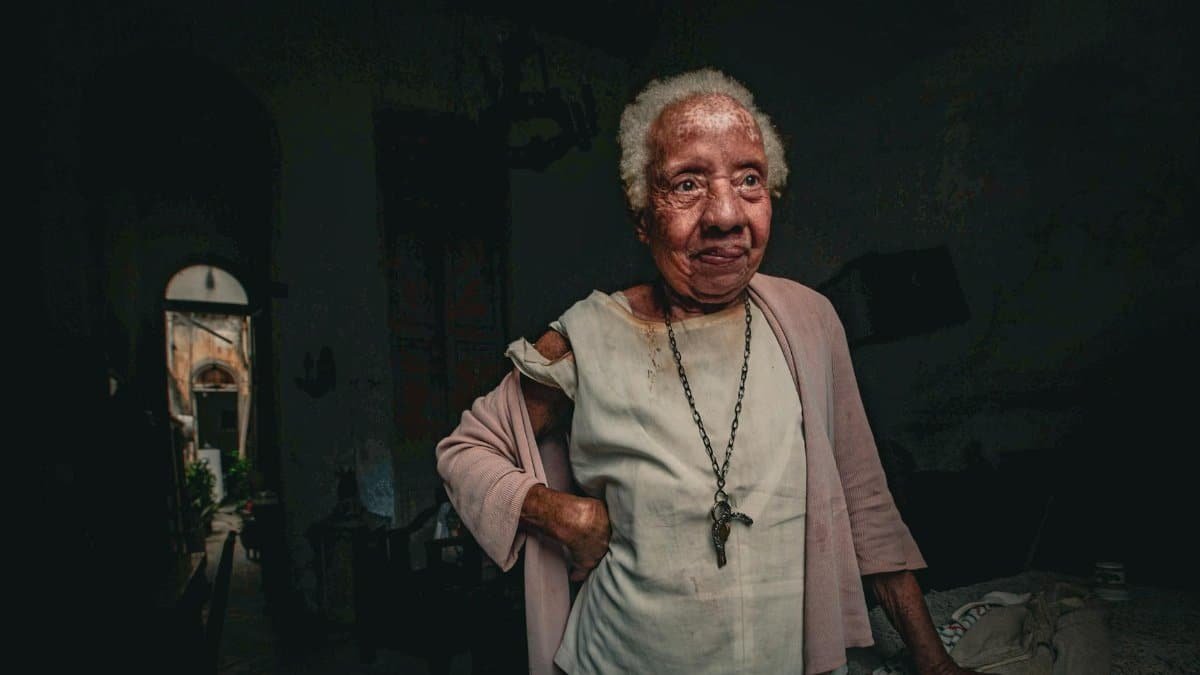 A senior woman with gray hair stands indoors, wearing a necklace, evoking a contemplative mood.
