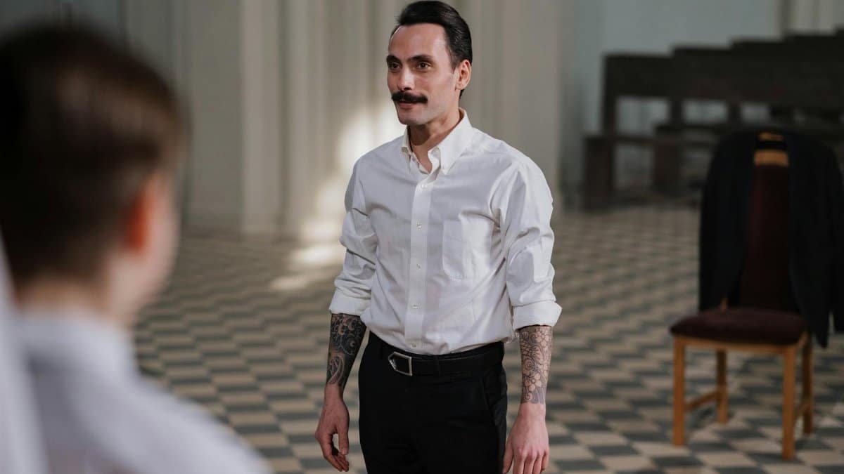 A man with tattoos and a moustache leads a practice indoors, wearing a white shirt.