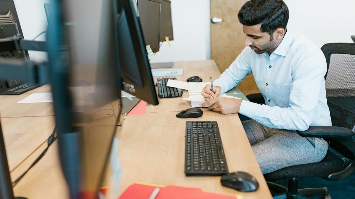 Business professional writing notes at desk with multiple monitors in a modern office setting.
