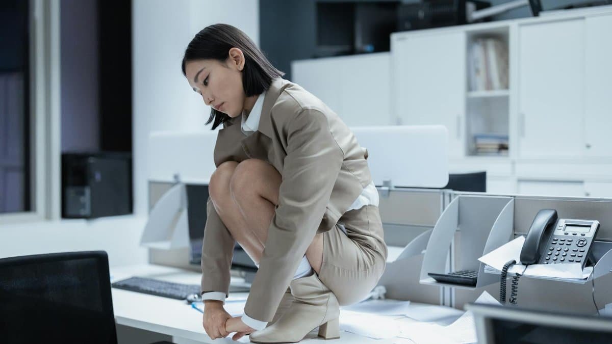 Asian businesswoman crouching on a desk in an office, depicting stress and overwork.