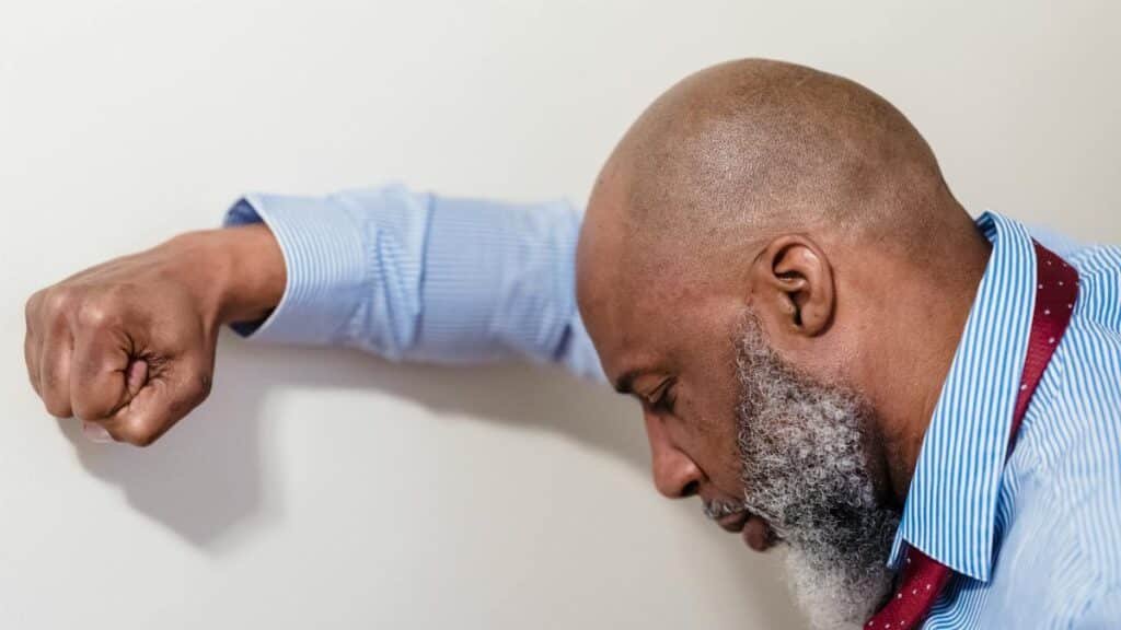 Bald man in blue shirt looks stressed, leaning against a white wall.