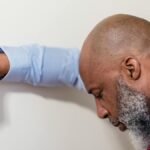 Bald man in blue shirt looks stressed, leaning against a white wall.