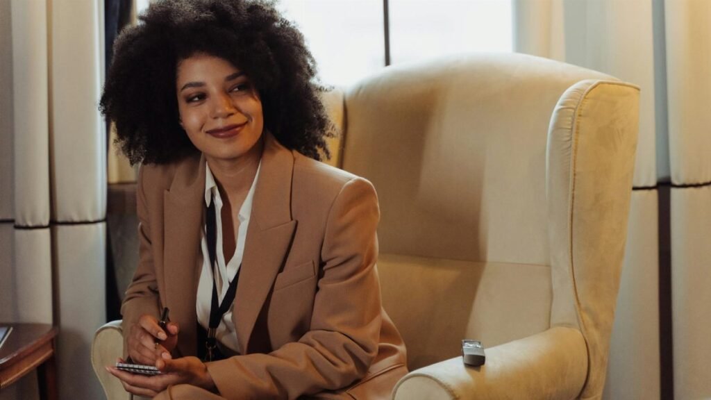 A confident woman in a brown blazer sits in an interview setting, taking notes.
