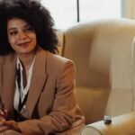 A confident woman in a brown blazer sits in an interview setting, taking notes.