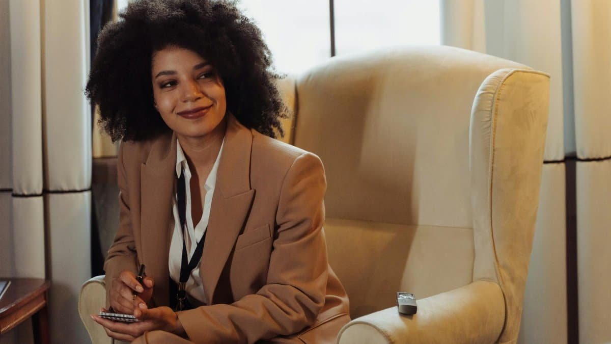 A confident woman in a brown blazer sits in an interview setting, taking notes.