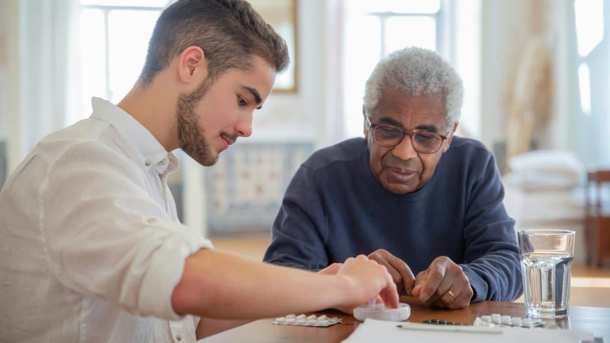A young volunteer helps an elderly man manage his medication at a nursing home.
