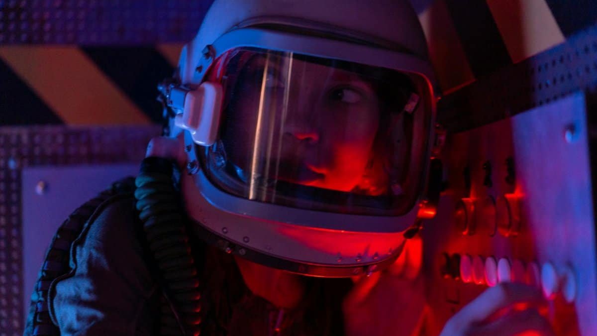 A woman astronaut wearing a spacesuit in a control room lit by red lights, looks focused.