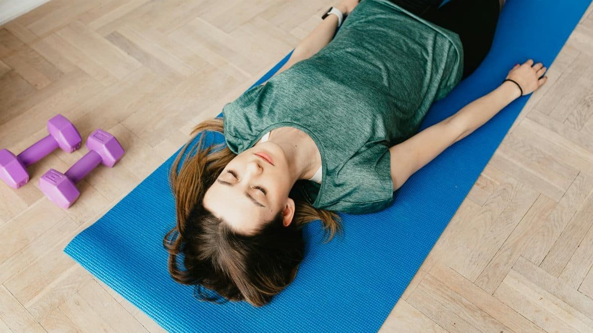 From above of young slender lady in casual clothes and wristwatch resting with closed eyes on blue yoga mat near dumbbells on floor in flat