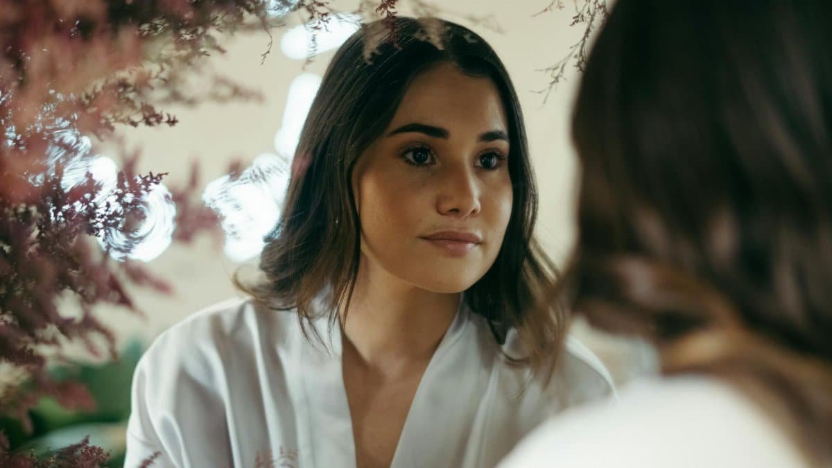 A woman gazes into a mirror surrounded by soft pink flowers, creating a serene and introspective ambiance.
