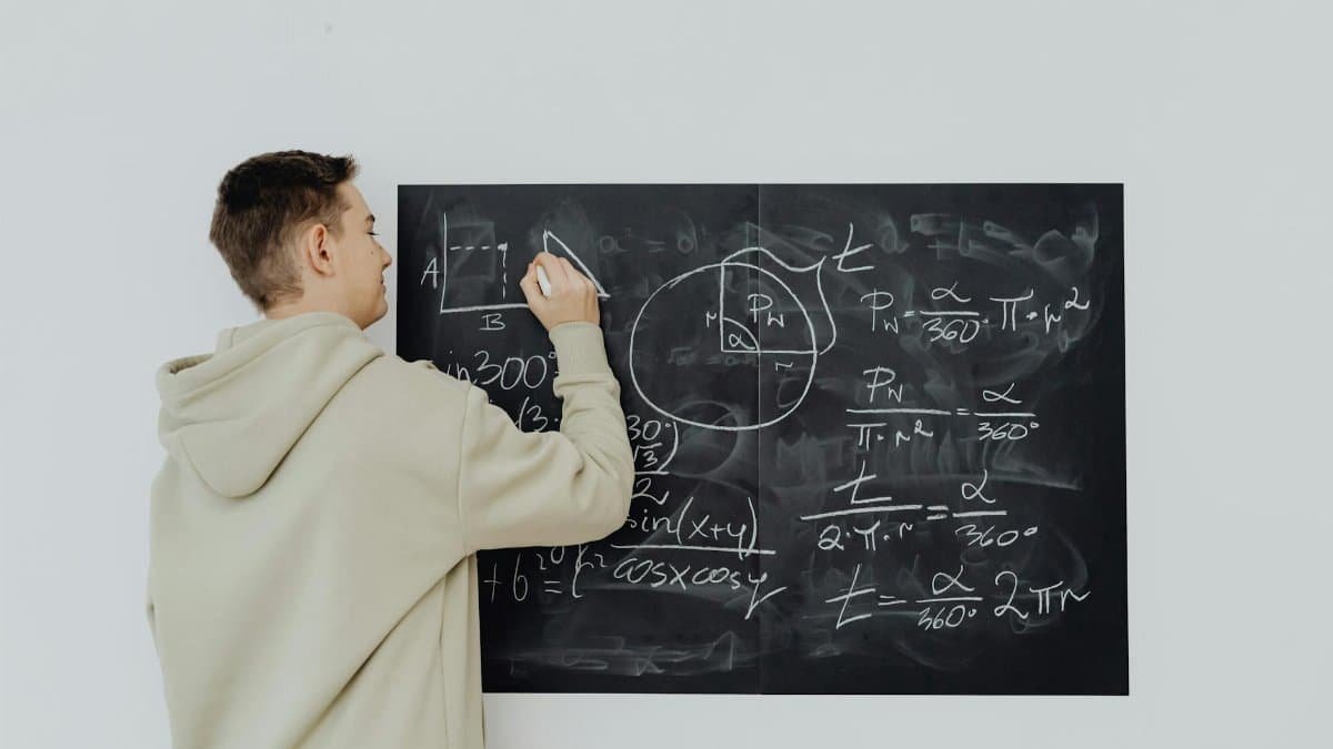 Teen boy in hoodie writes math formulas on a chalkboard, focused on solving algebra equations.