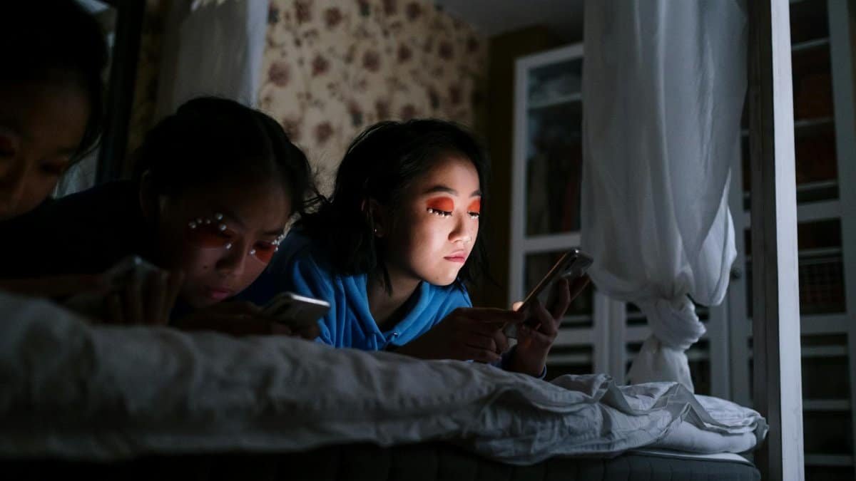 Three teenage girls using smartphones in a cozy bedroom setting, illuminated by screen light at night.