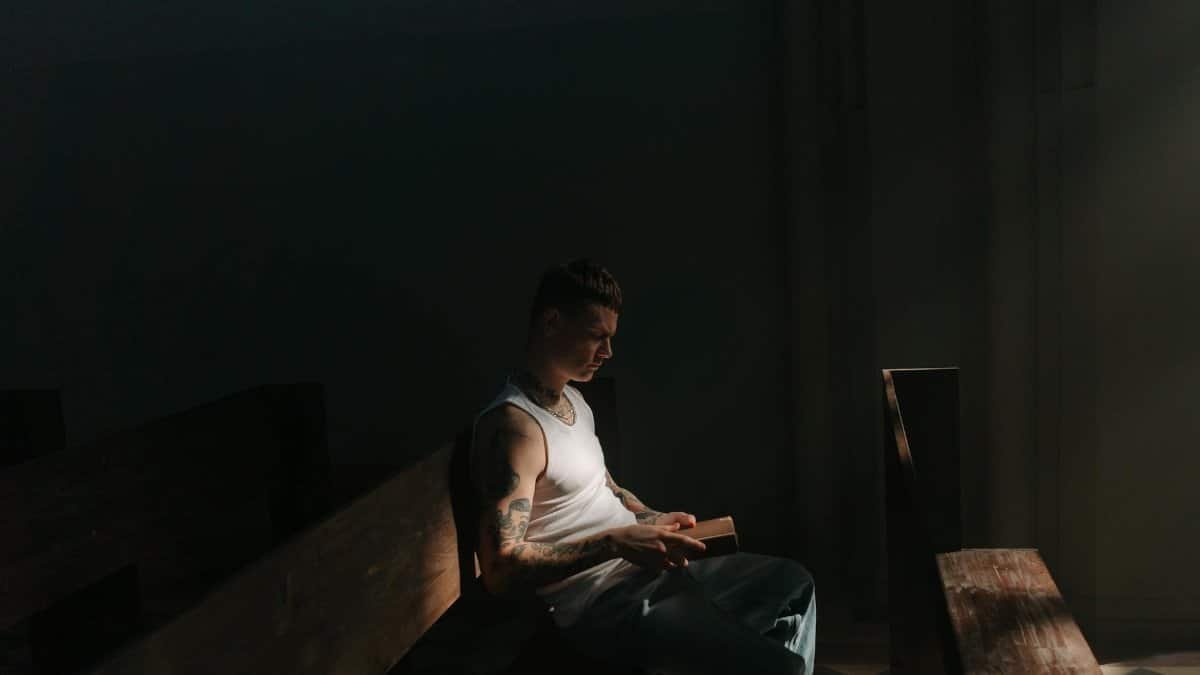 A tattooed man in a white tank top reads a book in dim lighting inside a church.