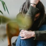 Anonymous young sorrowed female in casual outfit touching dark hair and embracing knees while sitting on chair at home