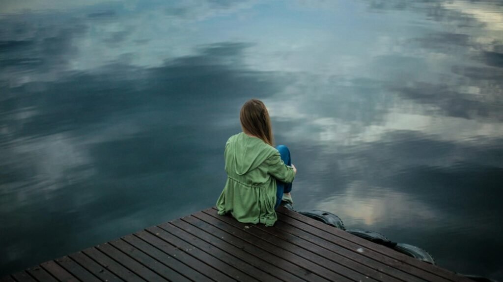 A woman sits on a wooden dock, reflecting by a calm lake under a cloudy sky.