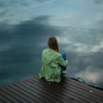 A woman sits on a wooden dock, reflecting by a calm lake under a cloudy sky.