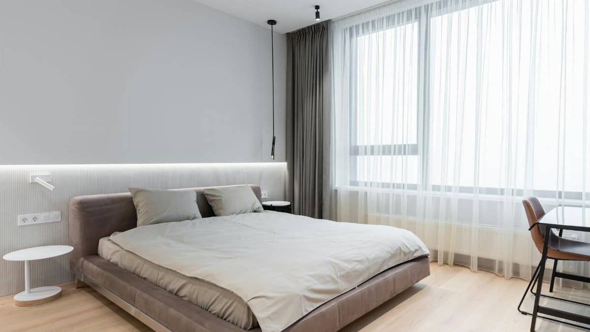 Interior of bright modern apartment with bed next to table with chair near windows with curtains