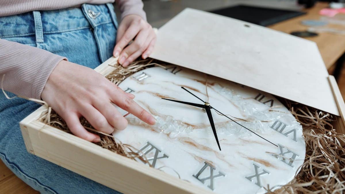 Hands gently inspecting a resin wall clock with Roman numerals in a wooden box, highlighting craftsmanship.