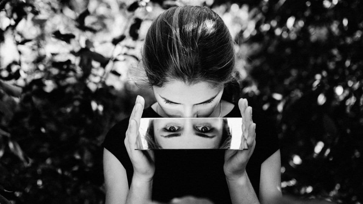 Black and white portrait of a woman gazing into a mirror outdoors, capturing reflection.