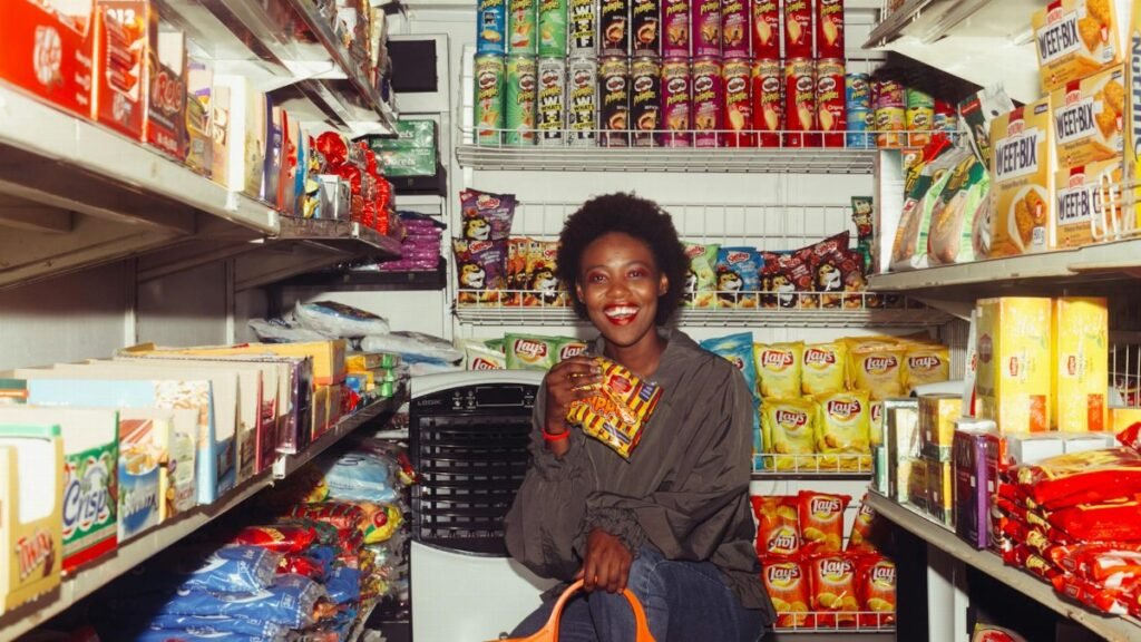 Happy black female in casual clothes with shopping bag demonstrating product squatting among grocery shelves while choosing goods in supermarket
