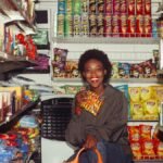 Happy black female in casual clothes with shopping bag demonstrating product squatting among grocery shelves while choosing goods in supermarket