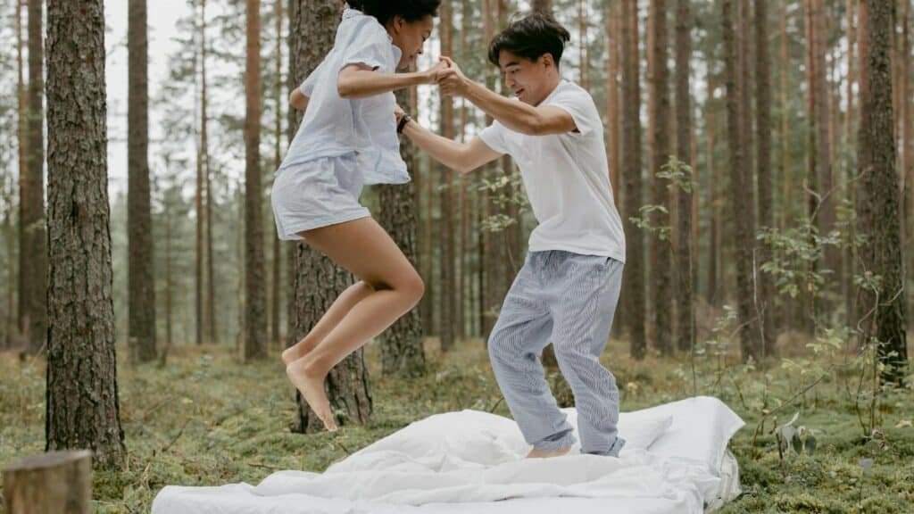 A joyful couple jumping on a bed outdoors in a peaceful forest setting.