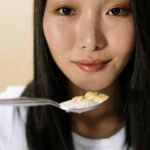 Portrait of an Asian woman holding a spoonful of cereal indoors, capturing a candid breakfast moment.
