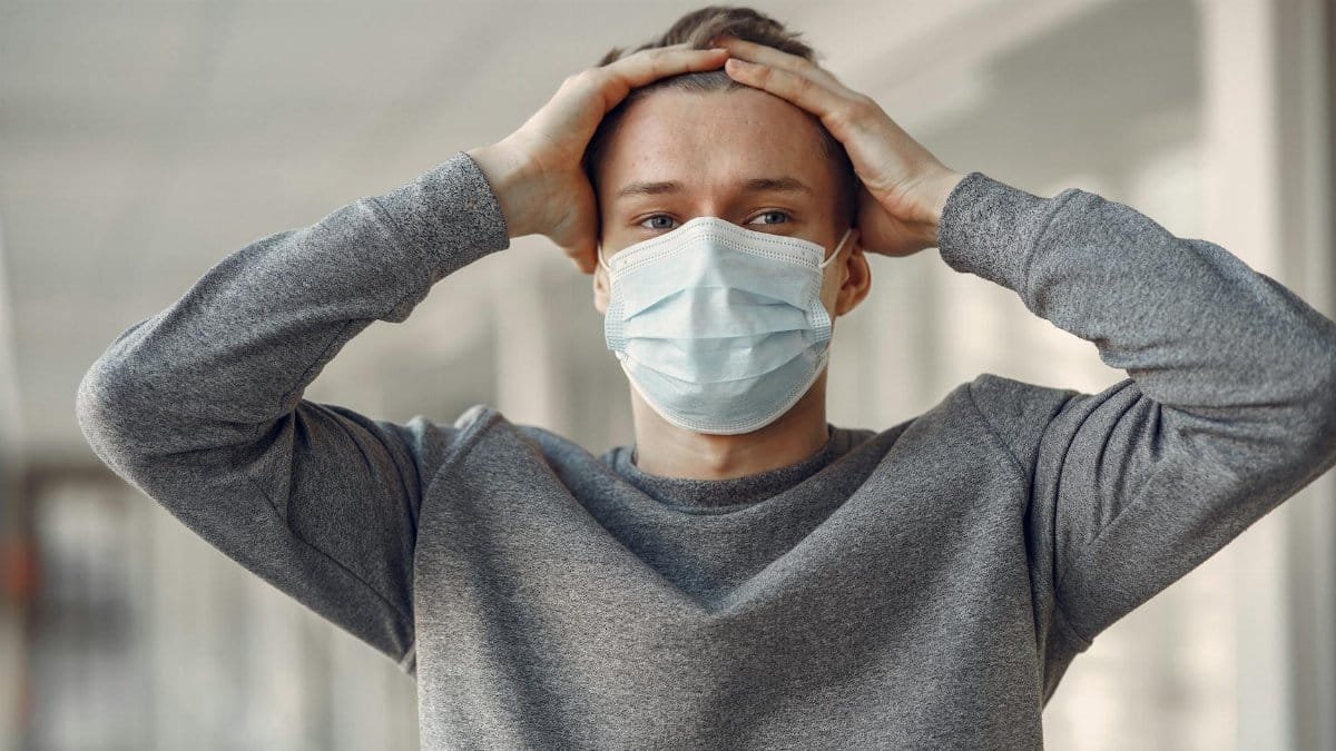 A young man wearing a medical face mask holds his head in anxiety indoors, reflecting stress and pandemic concerns.