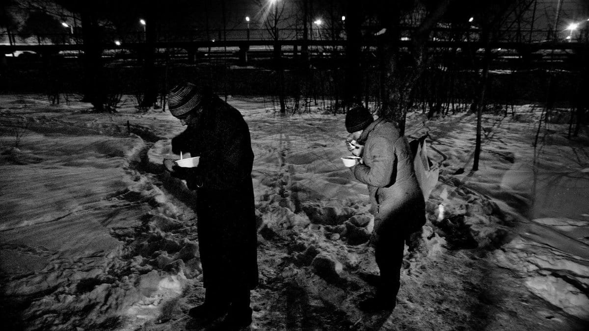 Two individuals standing and eating in a snowy urban setting at night.