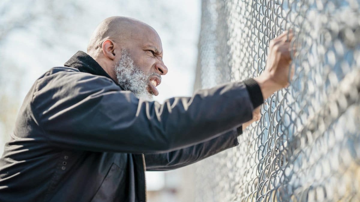 A frustrated man passionately grips a chain-link fence, expressing stress and determination.