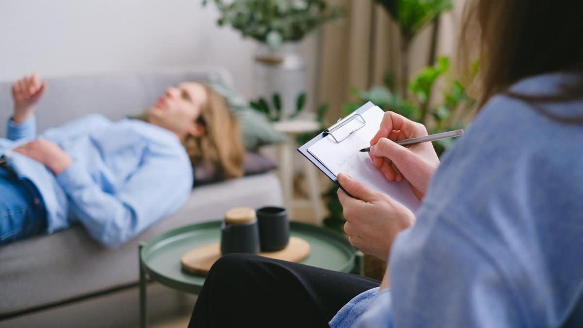 A therapy session in progress with a therapist taking notes as a patient reclines on a sofa.