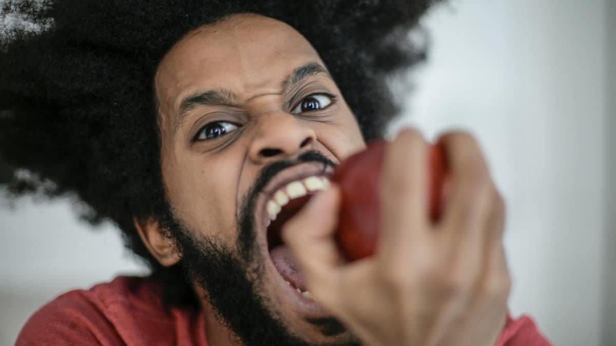 Energetic portrait of a man with afro hair biting into a red apple with an intense expression.